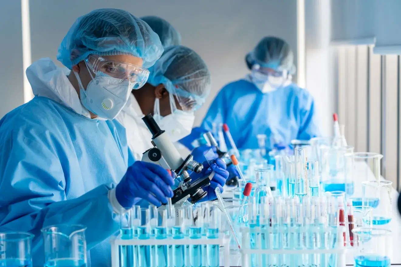 Three scientists working in a laboratory. Two are wearing a blue hairnet, safety glasses, white facemask, blue labcoat and blue gloves. One is wearing a blue hairnet, safety glasses, white facemask, and a white labcoat. A microscope and glassware are on a table in front of them.