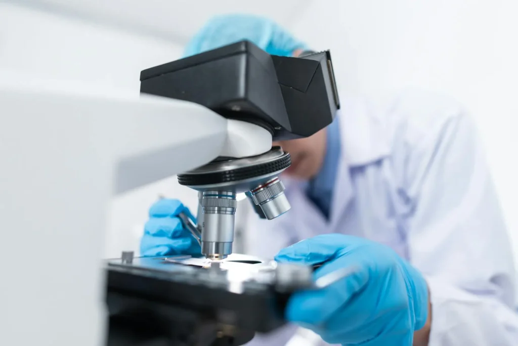 A scientist in a laboratory looking into a microscope, wearing a blue hairnet, white labcoat, and blue gloves