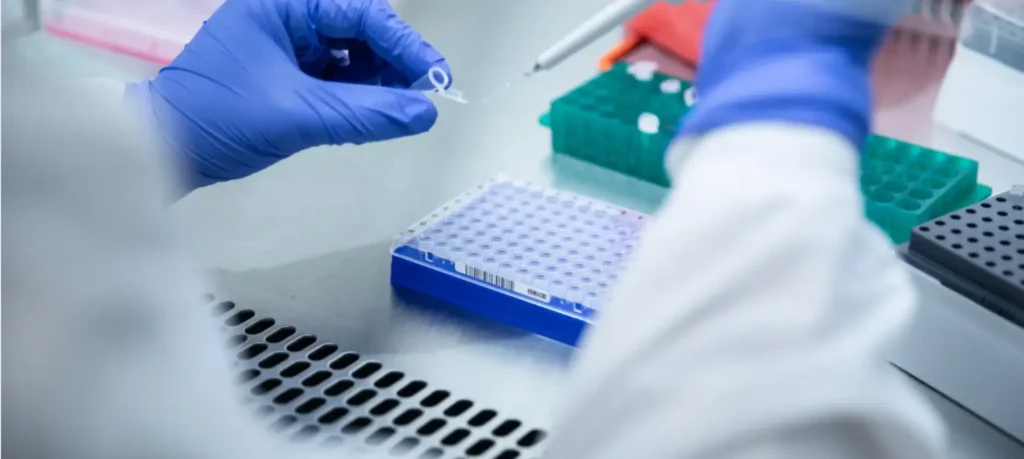 Gloved hands pipetting a liquid into a sample tube, above several multi-colored sample tube trays