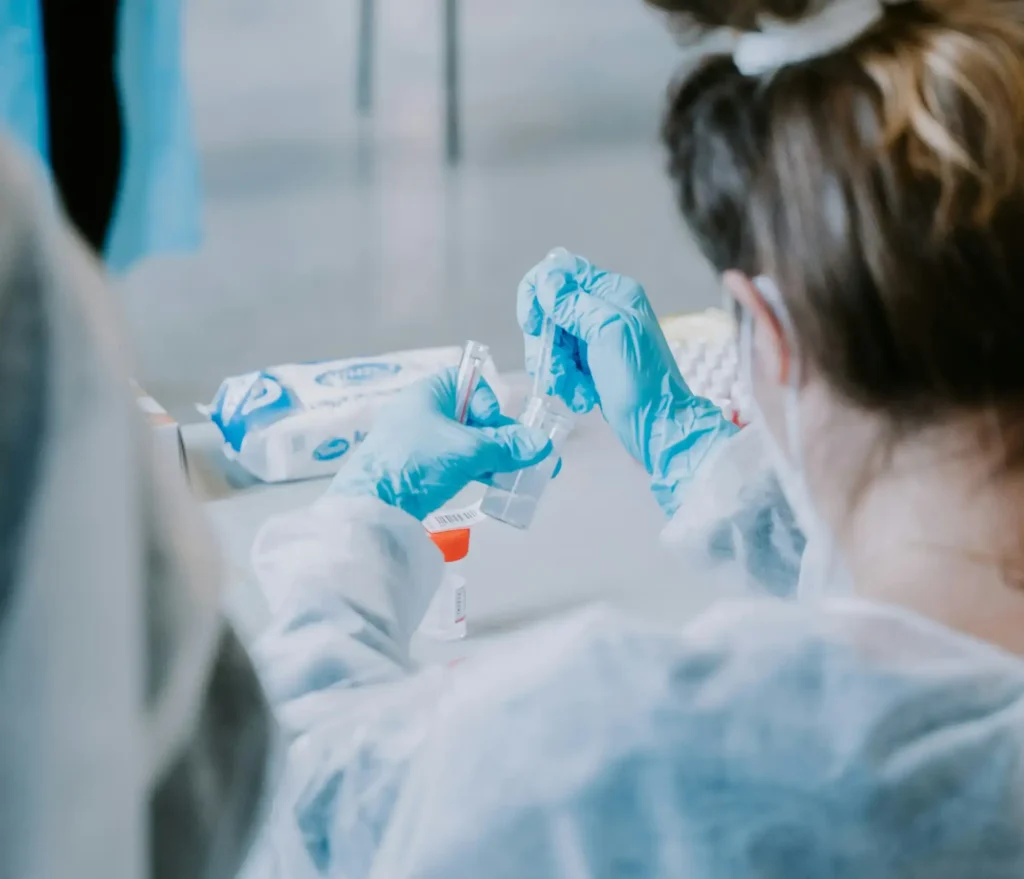 A scientist wearing a face covering, a white labcoat, and blue gloves inserting a swab into a small bottle.