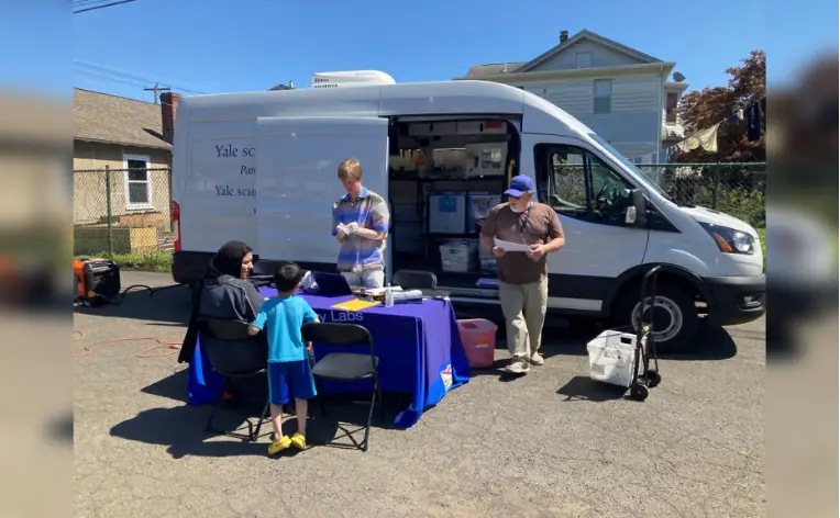Two men standing outside behind a table beside the opened side door of a white utility van. A seated woman and a standing child are in front of the table. The two men are workers assisting the woman and child.