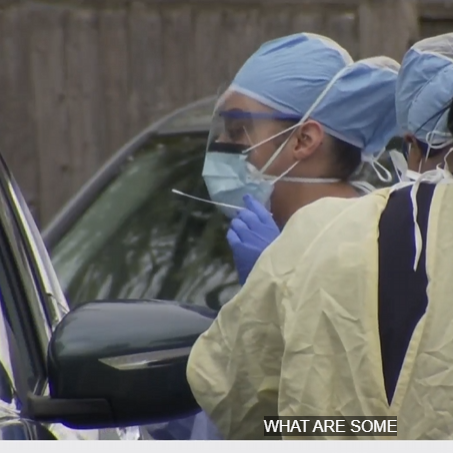 A woman wearing a blue head cover, blue face covering, clear face shield, safety glasses, and blue gloves holding a swab in her left hand.