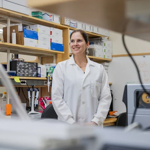 Anne Wyylie wearing a white labcoat standing in front a several shelves of laboratory boxes.