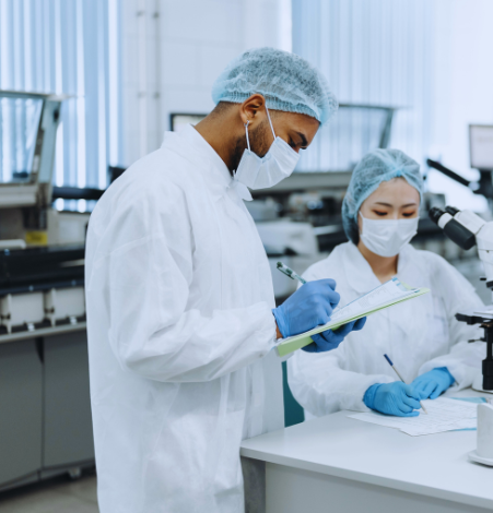 A man and woman standing in a laboratory wearing a blue hairnet, white face covering, white labcoat and blue gloves, both writing in a document.