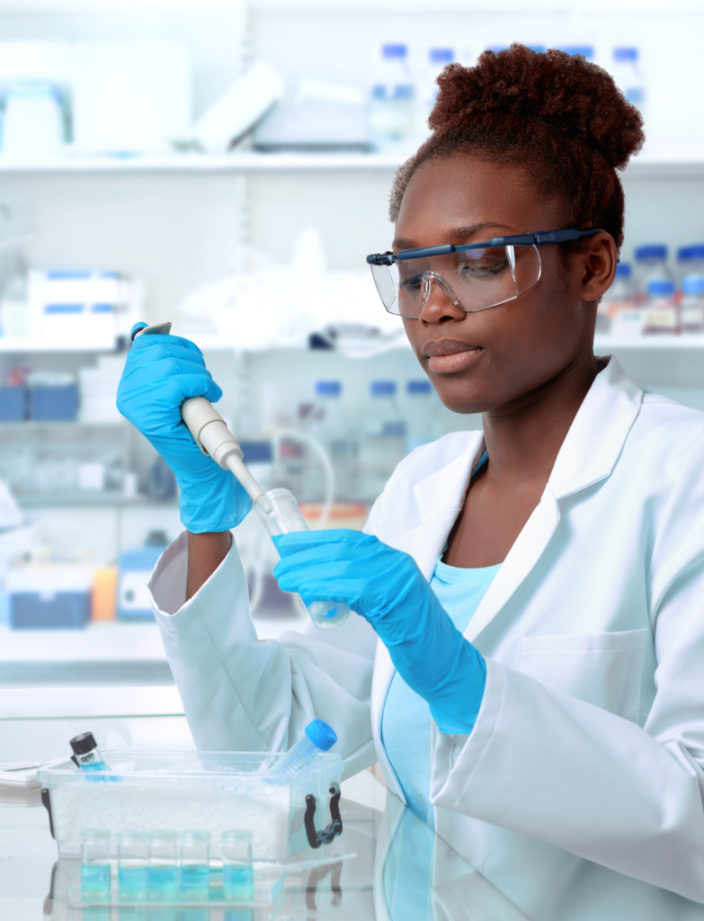 A woman scientist in a laboratory wearing safety glasses, a white labcoat, and blue gloves pipetting into a container. The pipette is in her right hand.