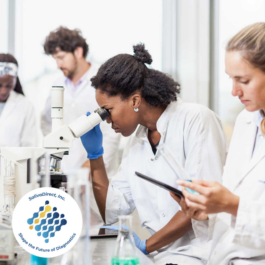A woman in a laboratory wearing a white labcoat and blue gloves looking into a microscope, standing beside another woman in a white labcoat staring at a computer tablet. A man and woman are standing in the background.